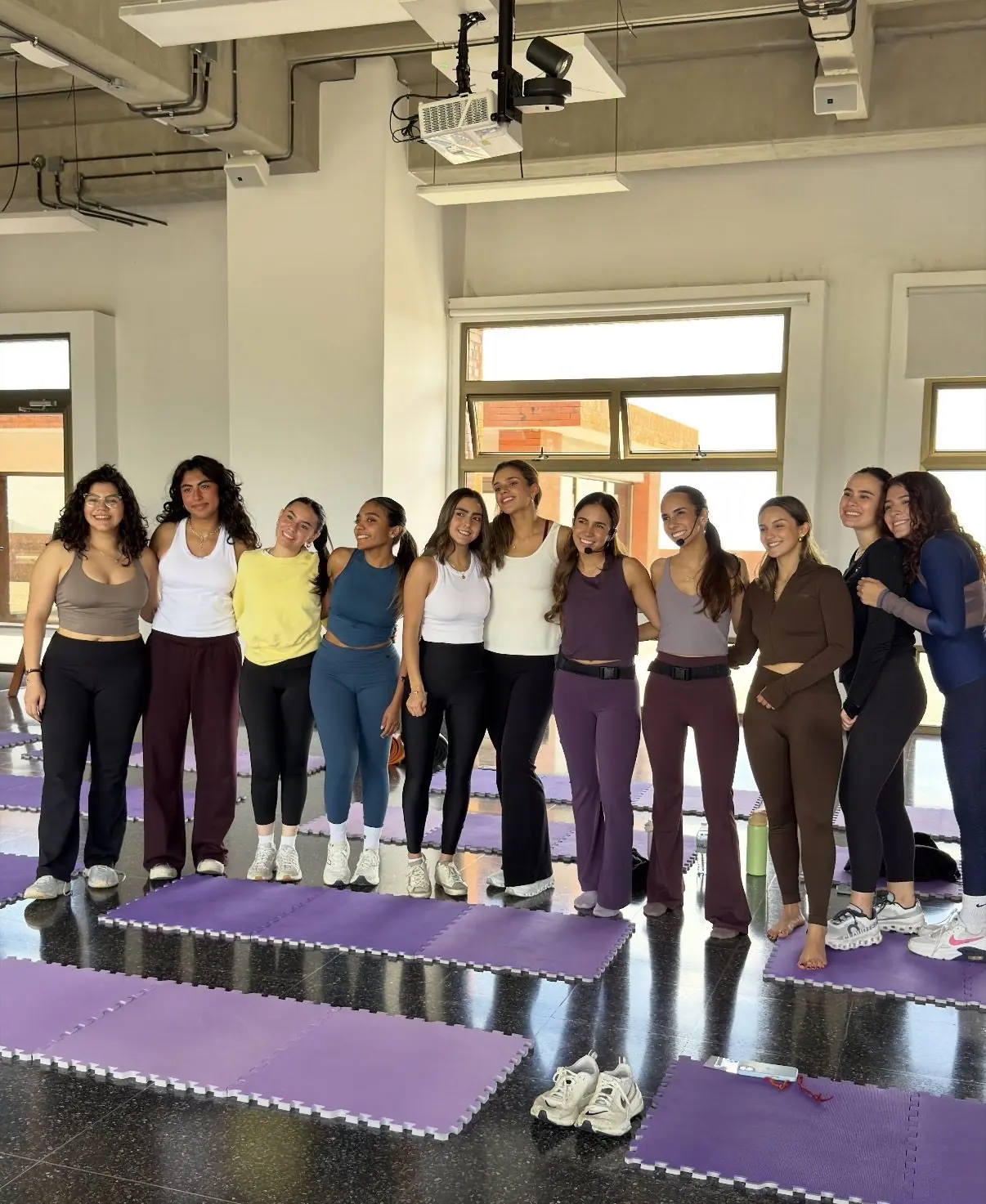 Foto grupal después de una clase en un espacio de oficina con piso negro brillante, colchonetas de foam moradas encajables, ventanales y techo con ductos y proyector a la vista