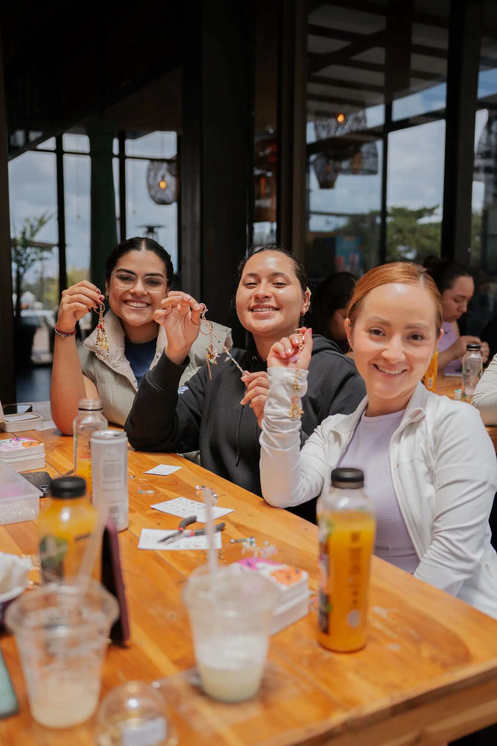 Tres asistentes sentadas a la mesa del taller muestran sonrientes los charms dorados que armaron, con jugos y vasos en primer plano