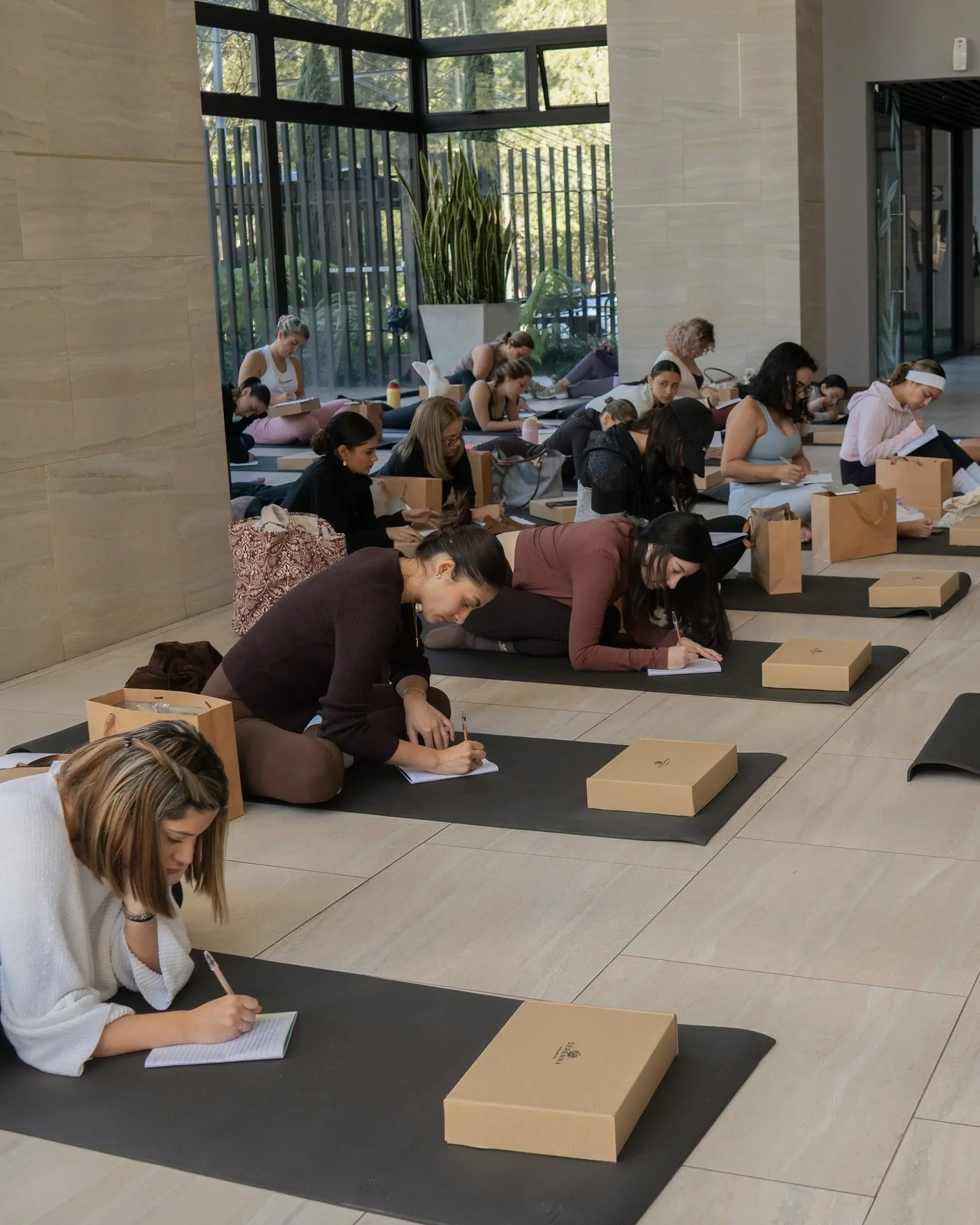 Fila larga de mujeres sentadas o recostadas en mats negros, escribiendo en sus journals dentro de un lobby con muros de piedra clara y ventanal al fondo