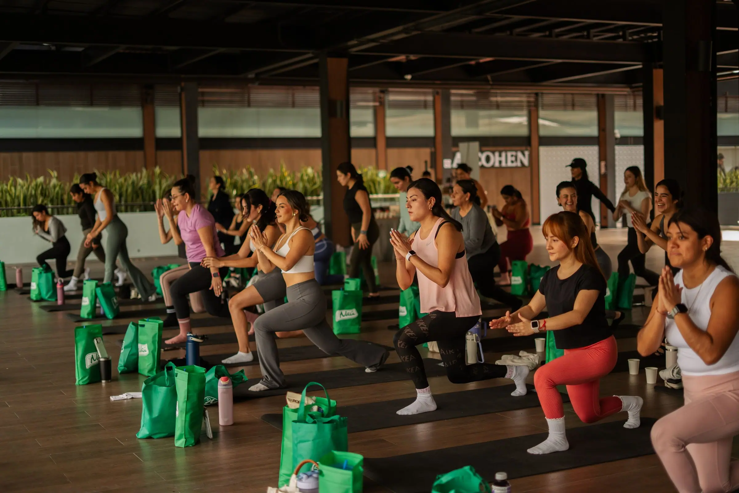 Clase masiva bajo techo abierto de centro comercial: filas de mujeres en estocada baja con las manos en posición de rezo, sonriendo